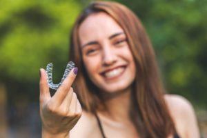 Blurry green background of a woman with brown hair smiling while she holds a clear aligner tray in the foreground